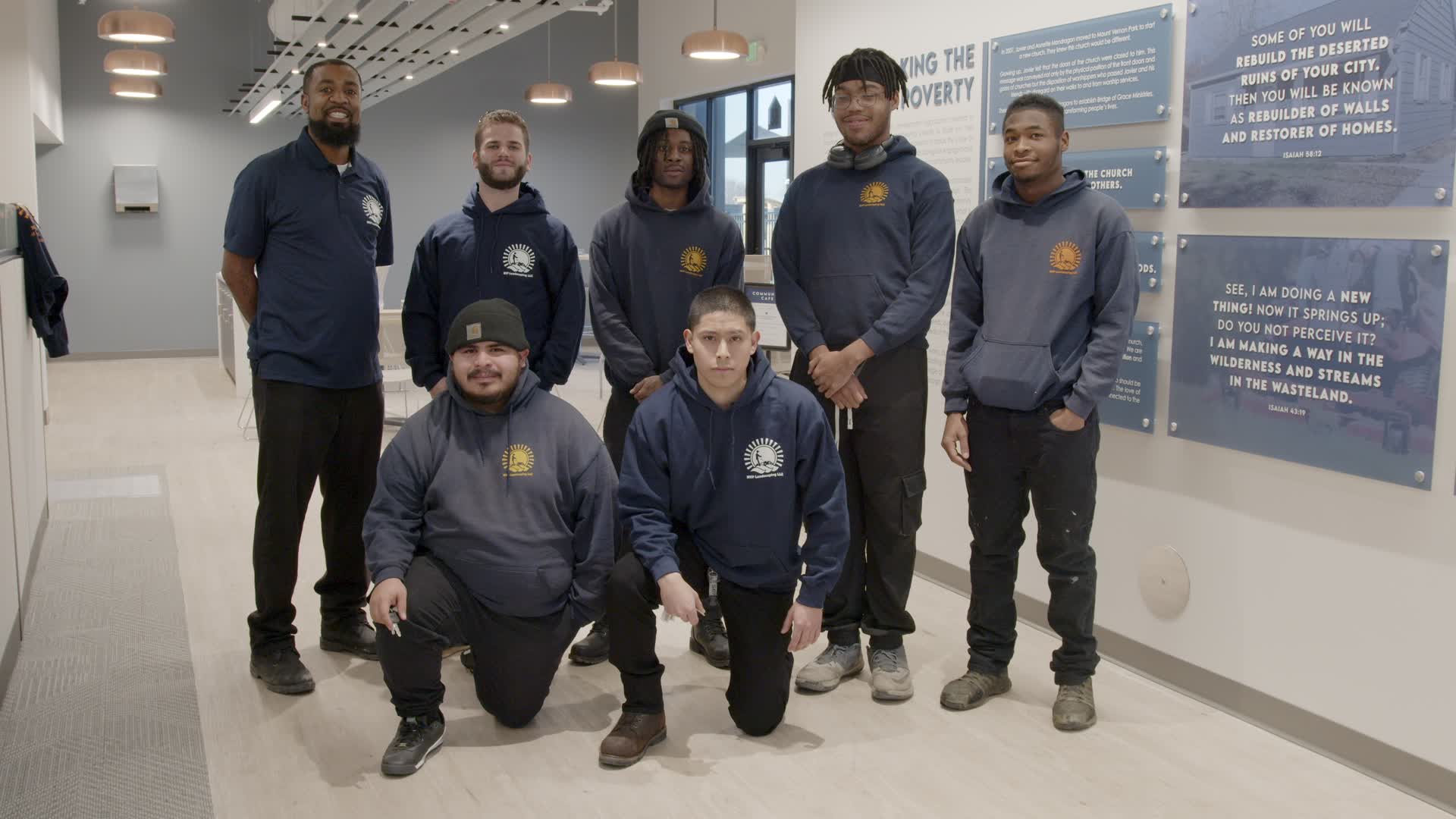 Seven men in navy hoodies pose for a group photo in a bright hallway, with two kneeling in front and blue wall plaques in the background.