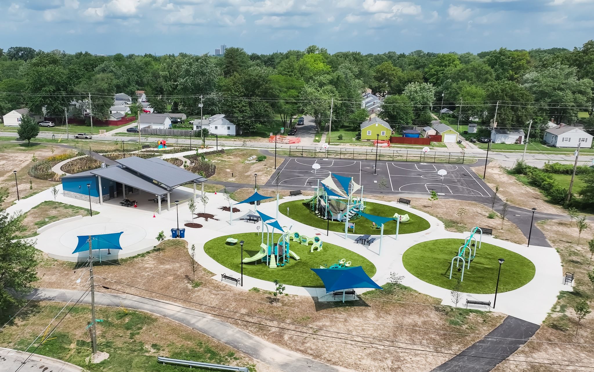 Aerial view of a colorful playground with blue shade sails, circular turf areas, slides, and a basketball court beside a neighborhood street.