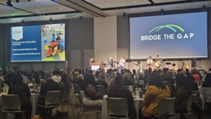Auditorium scene with a band performing on stage and a large screen reading'BRIDGE THE GAP' behind them.