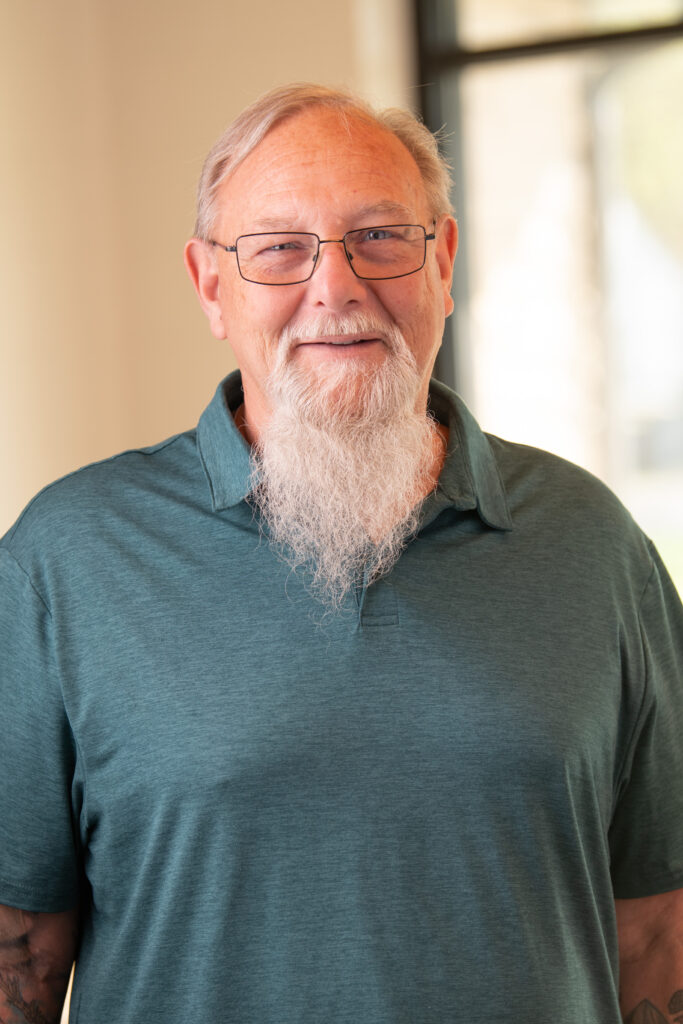 Smiling older man with glasses and a white beard wearing a teal polo, indoors by a window.
