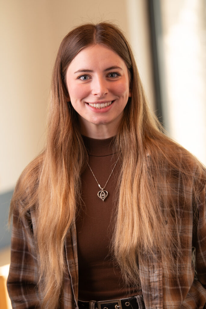 Young woman with long brown hair smiling, wearing a brown turtleneck and plaid shirt, indoors with soft lighting.