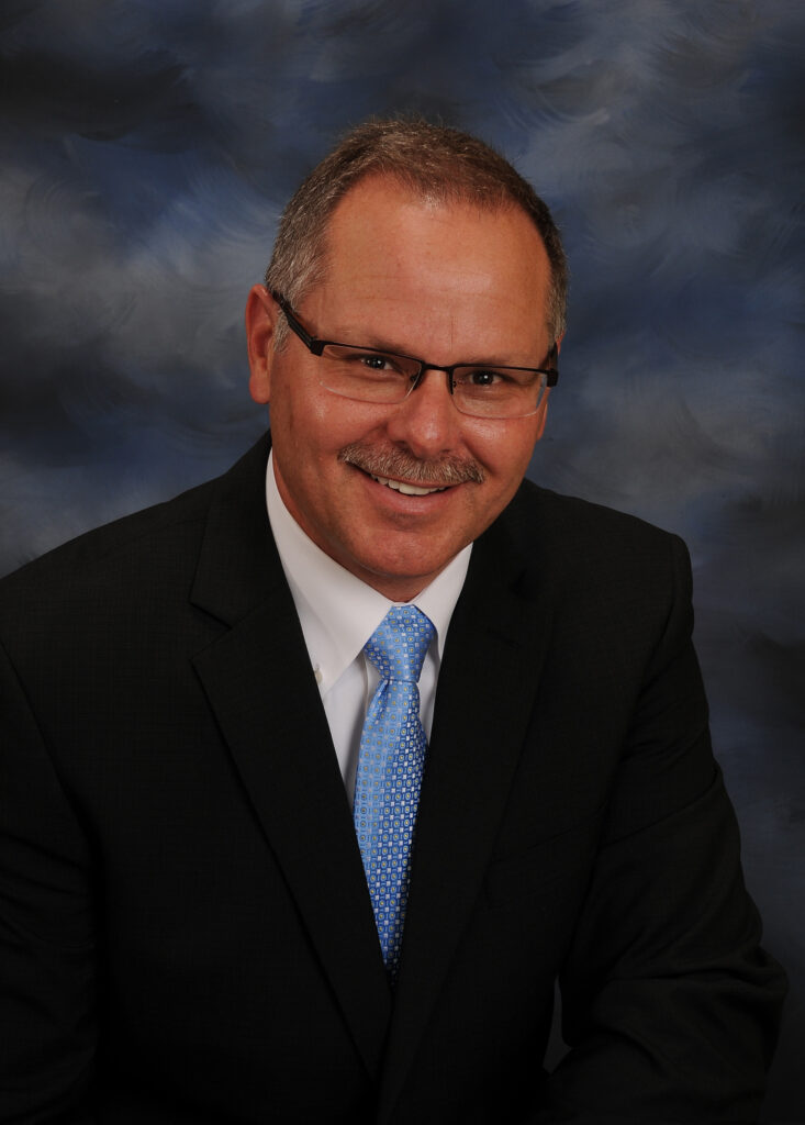 Professional portrait of a smiling middle-aged man in a dark suit, white shirt, and blue patterned tie, against a blue studio backdrop.
