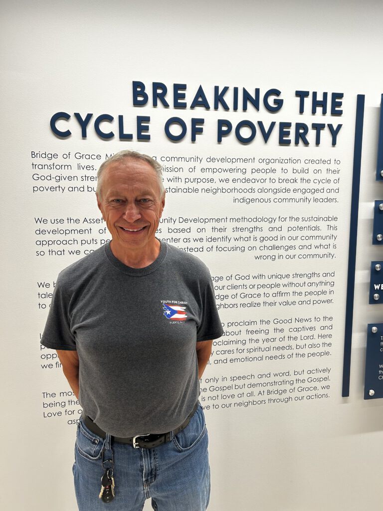 Man in a gray t-shirt and jeans smiles at camera while standing in front of a wall with large text reading 'BREAKING THE CYCLE OF POVERTY' and smaller paragraph text surrounding him.